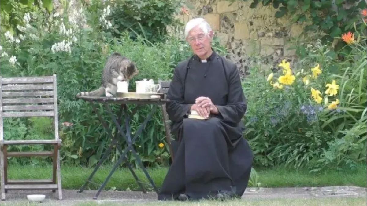 A cat sits on a table, sipping milk, the Dean of Canterbury sits beside him.